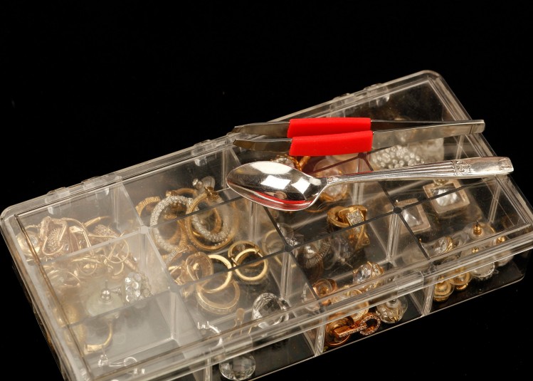 Close-up of the tweezers and demitasse spoon on top of the closed box of earrings. The earrings are visible through the clear plastic cover.