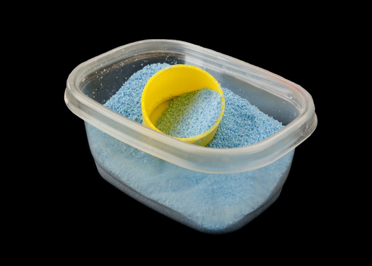 A studio shot of a rectangular plastic dish, normally used for food storage, containing 1-2 cups of powdered detergent. A small yellow measuring cup stands ready.