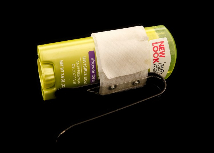 Studio shot of a yellow stick of roll-on deodorant,  with a holder wrapped around it. The holder consists of a white velcro strap wrapped around the stick, and a protruding piece of black metal that forms a loop and is attached to the velcro with screws. The metal loop holder comes out from the stick at an angle so the loop rests securely on Cindy's hand as she applies the deodorant.