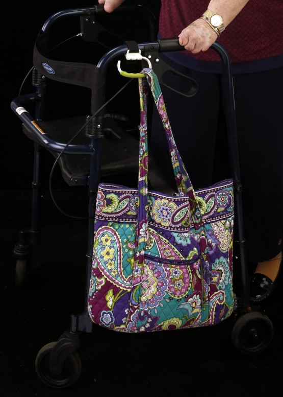 Studio close up of carabiner attached to the left handle of the walker, and angled down. It holds a paisley pastel-colored quilted cloth bag. Cindy'd hands are visible, holding the walker handles.