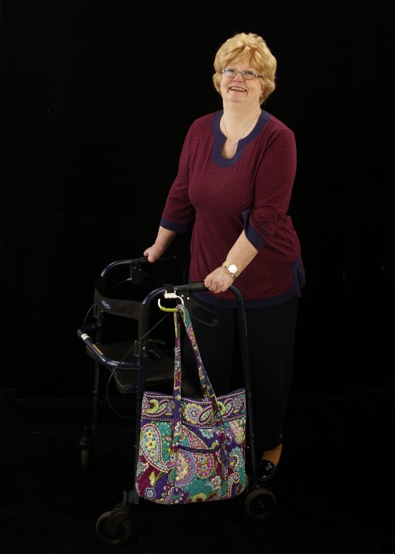 Studio shot of Cindy standing at her walker against a black background. Cindy is in a maroon shirt, is smiling at the camera, and has her two hands on the handles of the walker. The carabiner is attached to the left handle of the walker, and is angled down. It holds a paisley pastel-colored quilted cloth bag.