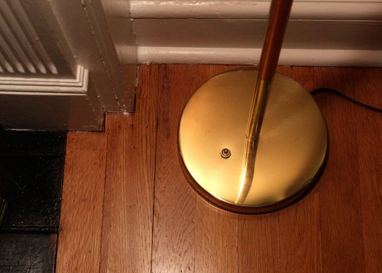 An interior shot of Cindy's floor lamp that's activated by a foot switch: a small brass button for stepping on and off of a large circular brass lamp base.
